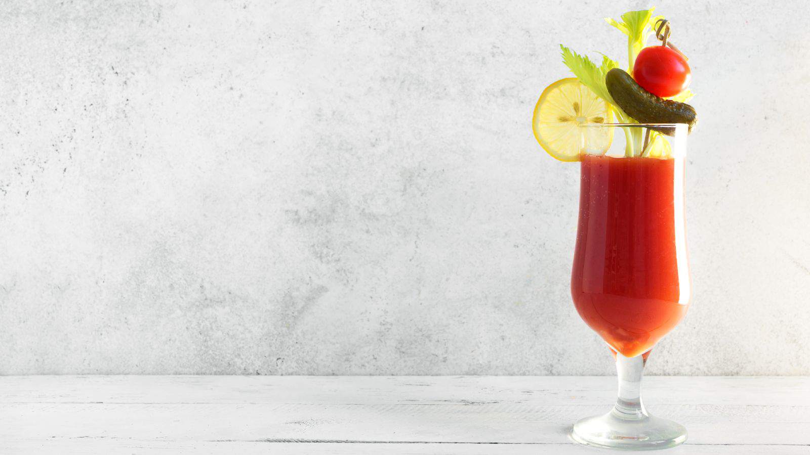A tall glass of Bloody Mary, garnished with celery, a lemon slice, a cherry tomato, and a pickle spear, standing against a light gray background.