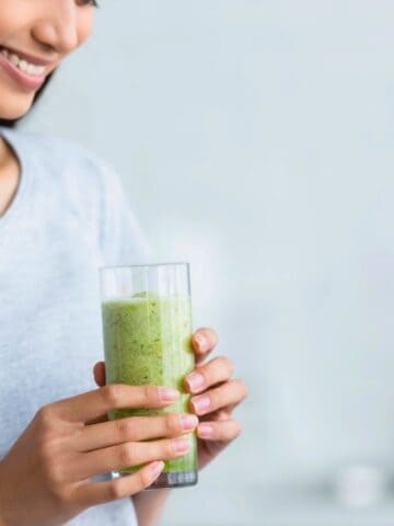 A person in a gray shirt holds a glass filled with a green smoothie. The background is softly blurred, suggesting a kitchen setting.