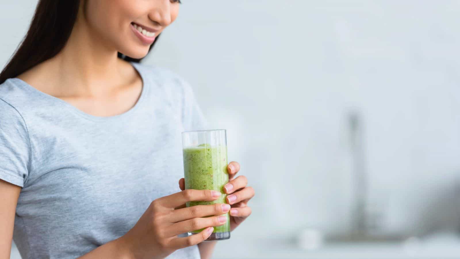 A person in a gray shirt holds a glass filled with a green smoothie. The background is softly blurred, suggesting a kitchen setting.