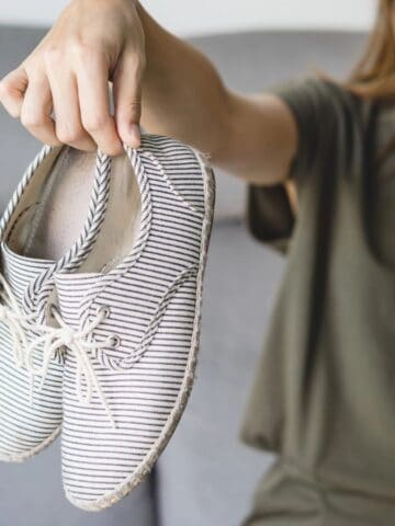 A person holds a pair of striped shoes at arm's length while pinching their nose, suggesting an unpleasant odor. They sit on a couch, perhaps pondering surprising coffee grounds uses to tackle the smell. The background is softly blurred.