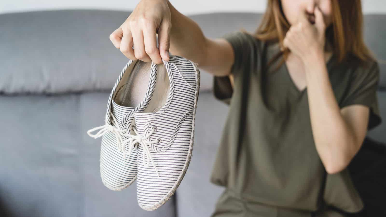 A person holds a pair of striped shoes at arm's length while pinching their nose, suggesting an unpleasant odor. They sit on a couch, perhaps pondering surprising coffee grounds uses to tackle the smell. The background is softly blurred.