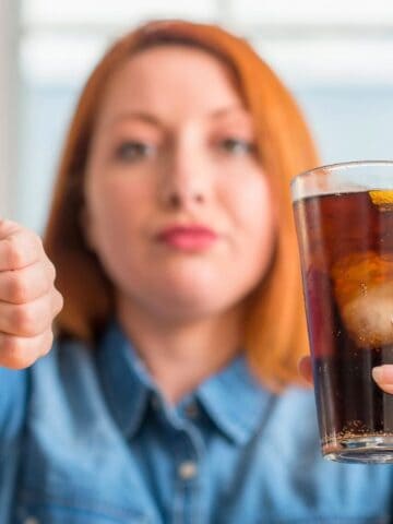 A person with red hair, wearing a denim shirt, holds a glass of dark soda with ice and lemon in one hand while giving a thumbs-down with the other hand. The expression is neutral and focused on the camera.