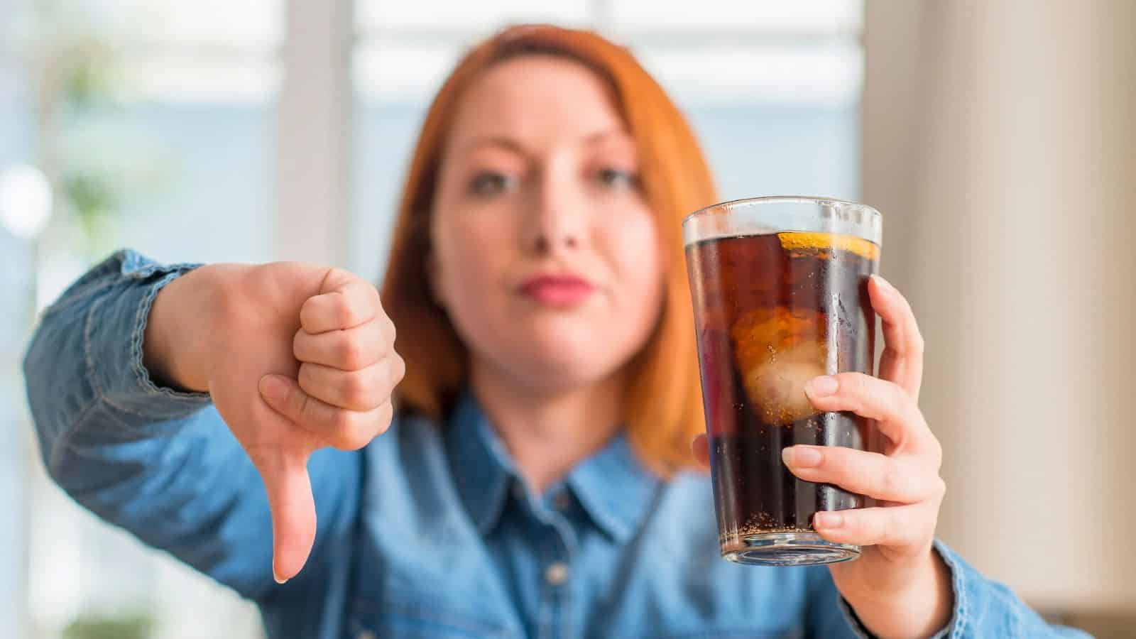 A person with red hair, wearing a denim shirt, holds a glass of dark soda with ice and lemon in one hand while giving a thumbs-down with the other hand. The expression is neutral and focused on the camera.