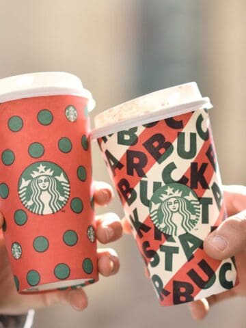 Two people holding Starbucks holiday-themed cups, one red with green polka dots and the other white with a bold logo pattern, in an outdoor setting. Both individuals are wearing jackets, suggesting cool weather.
