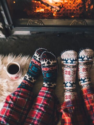 Two people wearing patterned socks rest their feet on a wooden surface covered with a furry blanket, facing a fireplace. Two mugs of coffee are placed beside their feet. They're wearing plaid pajamas. The scene is cozy and warm.