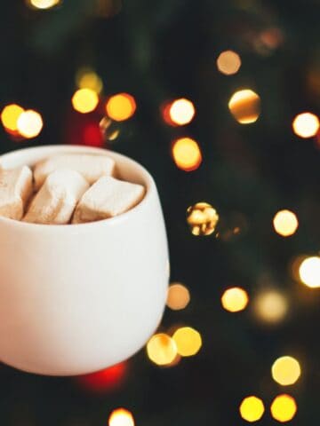 A hand holds a white mug filled with marshmallows, against a blurred background of festive, glowing lights.