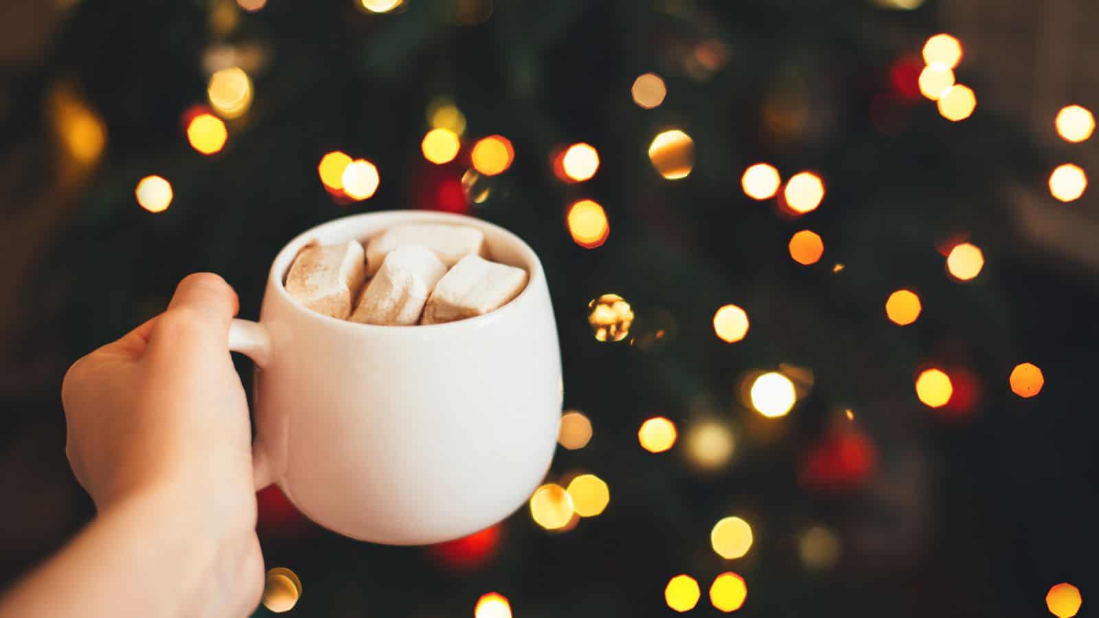 A hand holds a white mug filled with marshmallows, against a blurred background of festive, glowing lights.