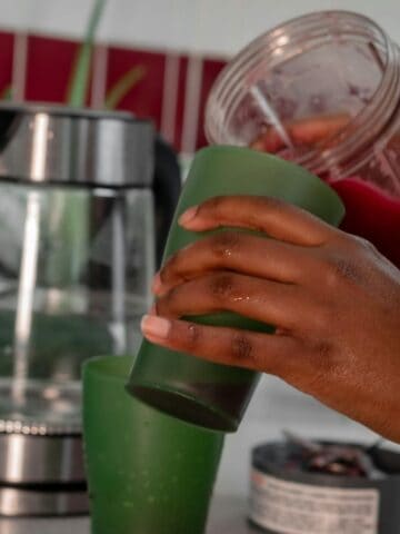 A person pours a red smoothie from a blender container into a green cup in a kitchen. A glass kettle and other kitchen items are in the background.