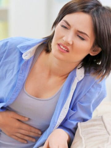 A woman in a blue shirt leans forward slightly with a pained expression, holding her stomach with one hand. She appears to be experiencing discomfort or pain. The background is softly blurred, suggesting an indoor setting.