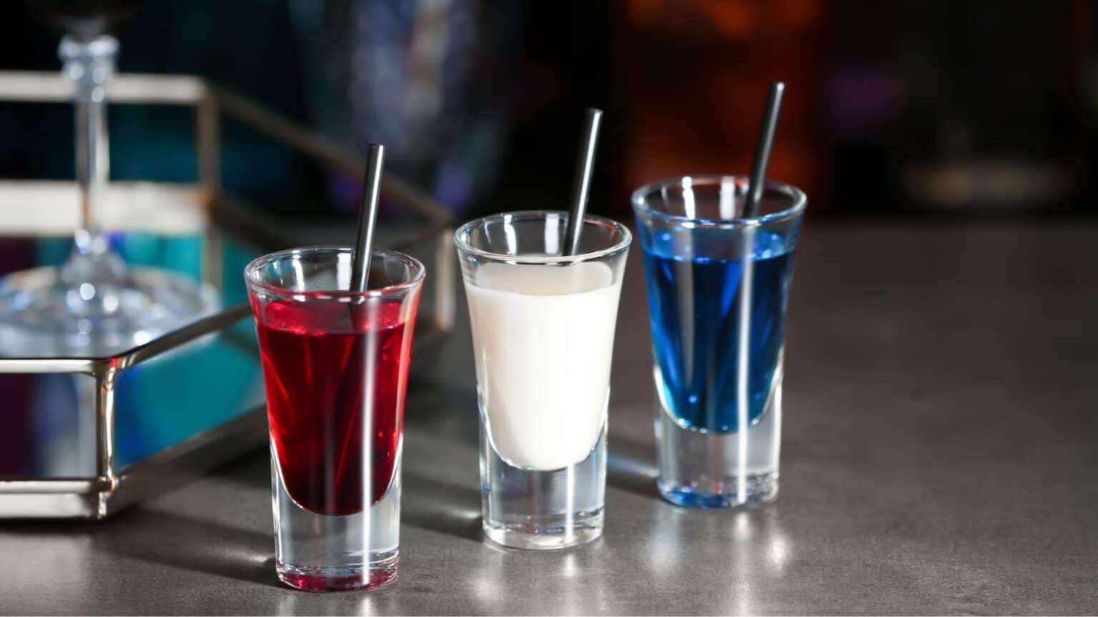 Three shot glasses are lined up on a counter, each containing a different brightly colored liquid: red, white, and blue. Black straws are placed in each glass. A metal tray with a wine glass is partially visible in the background.