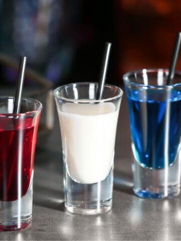 Three shot glasses are lined up on a counter, each containing a different brightly colored liquid: red, white, and blue. Black straws are placed in each glass. A metal tray with a wine glass is partially visible in the background.