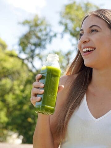 A young woman with long brown hair smiles while holding a bottle of green juice. She is wearing a white tank top and is standing outdoors on a sunny day with trees and a person in the background.