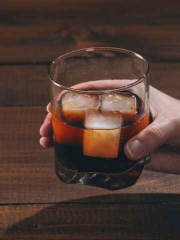 A hand holds a glass of dark liquid with ice cubes above a wooden table.
