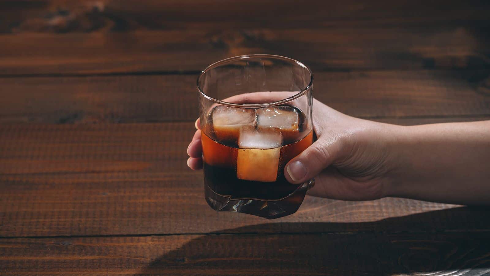 A hand holds a glass of dark liquid with ice cubes above a wooden table.
