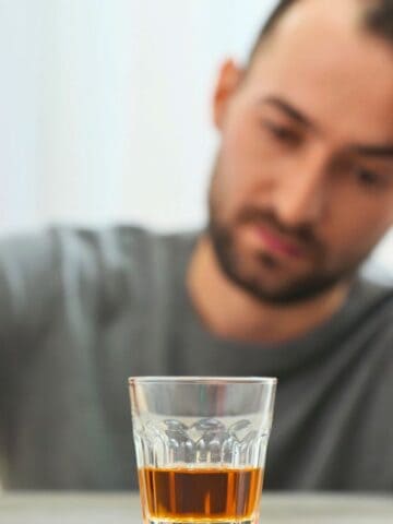 A man sits at a table holding a bottle of amber-colored liquor. In front of him, there is a glass filled with the same liquid. He rests his head on his hand, appearing contemplative or distressed.
