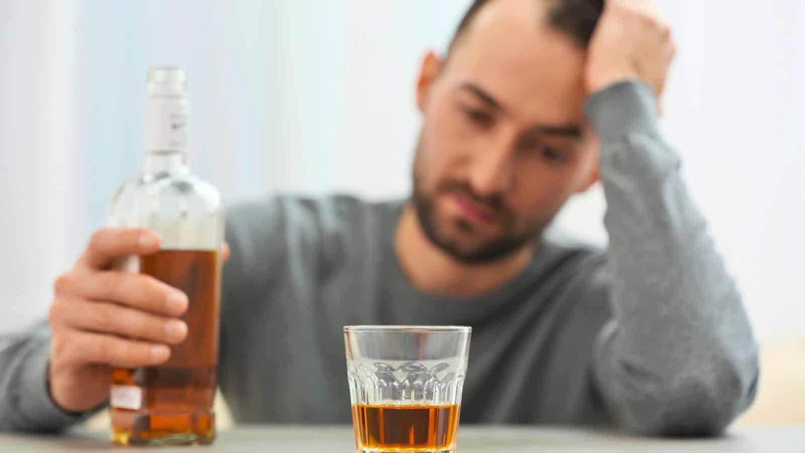 A man sits at a table holding a bottle of amber-colored liquor. In front of him, there is a glass filled with the same liquid. He rests his head on his hand, appearing contemplative or distressed.