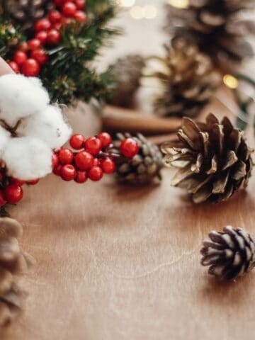 A hand arranges a holiday decoration with cotton, red berries, and evergreen branches. Pinecones and a ball of dark green yarn are scattered on a wooden surface. Twinkling lights are partially visible in the background.