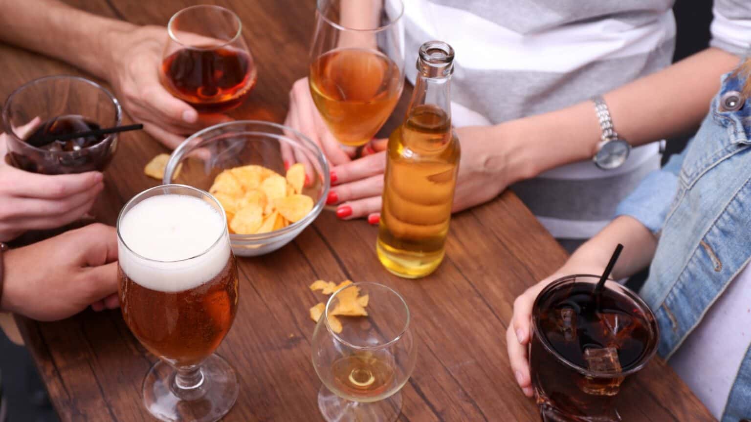 A group of people gathered around a wooden table with various drinks, including beer and cocktails, in glasses. A bowl of chips is placed in the center, and scattered chips are visible on the table.