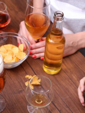 A group of people gathered around a wooden table with various drinks, including beer and cocktails, in glasses. A bowl of chips is placed in the center, and scattered chips are visible on the table.
