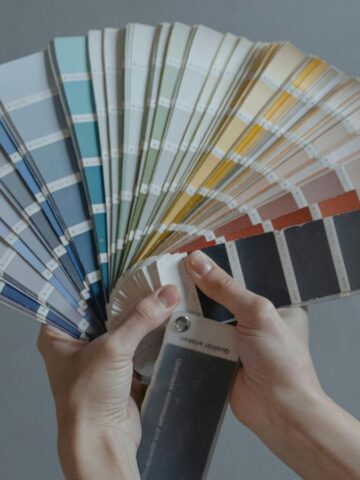 A person is holding a fanned-out set of color swatches against a plain gray background. The swatches display a gradient of colors ranging from blue and green to orange and black.