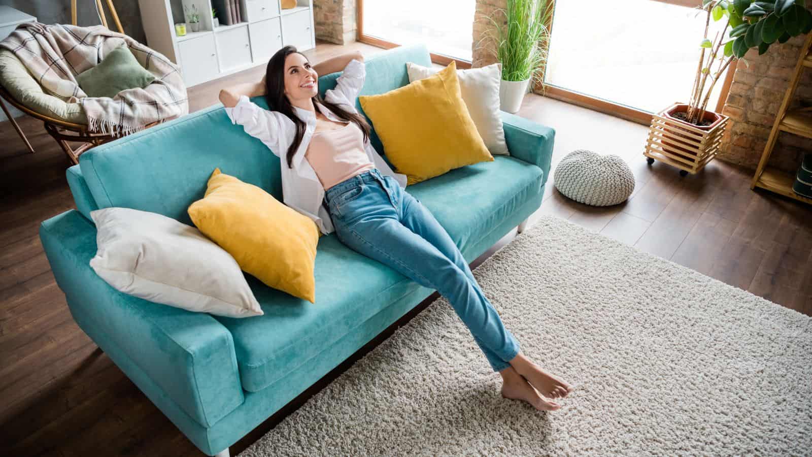 A woman relaxes on a turquoise sofa with her arms behind her head. She is surrounded by yellow and white cushions. The room has wooden floors, large windows, and potted plants, creating a bright and cozy atmosphere.