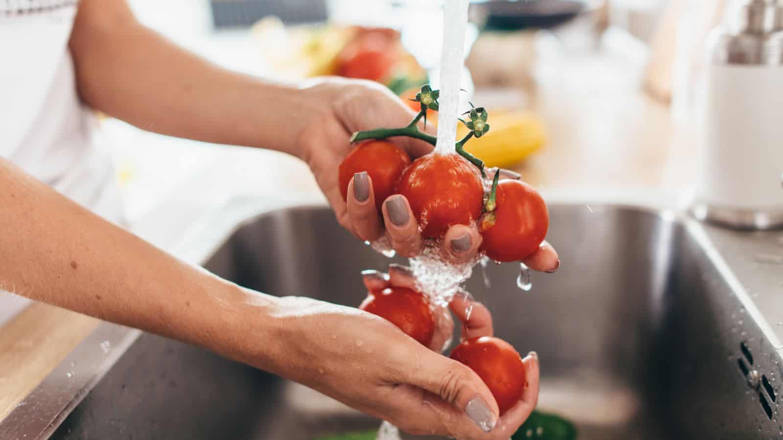 A person is washing a bunch of red tomatoes under running water in a kitchen sink. Both hands are holding the tomatoes, and droplets of water are visible. The background includes a blurred view of other kitchen items.