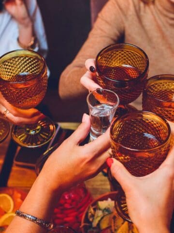 A group of people toasting with amber-colored glasses filled with drinks. The table is set with various snacks and colorful food items. Hands are visible, holding up the glasses in a celebratory gesture.