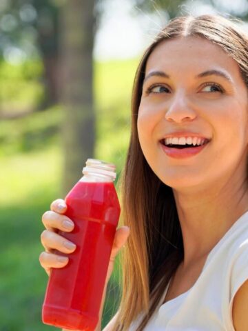 A person with long hair and a white shirt is smiling while holding a bottle of red juice. They are outdoors with green grass and trees in the background, suggesting a park setting.