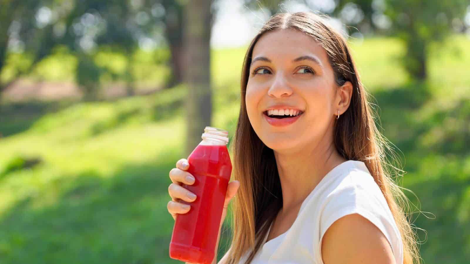 A person with long hair and a white shirt is smiling while holding a bottle of red juice. They are outdoors with green grass and trees in the background, suggesting a park setting.