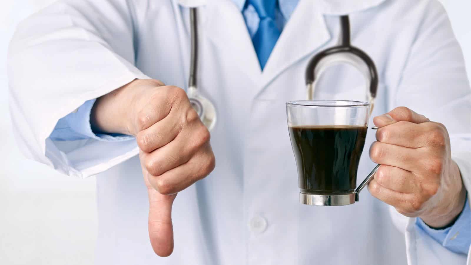 A person in a white lab coat with a stethoscope shows a thumbs-down gesture while holding a glass of dark coffee.