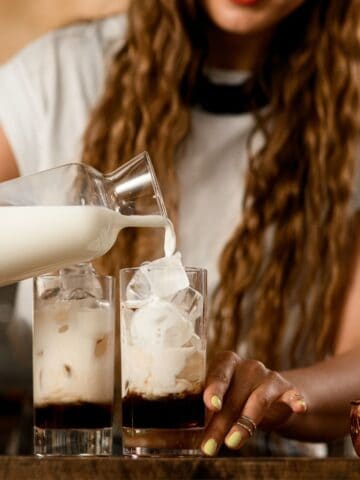 A person with long hair is pouring milk into two tall glasses of iced coffee, illustrating tips for perfect iced coffee. A copper mug sits on the counter nearby, adding charm to the cozy café atmosphere.
