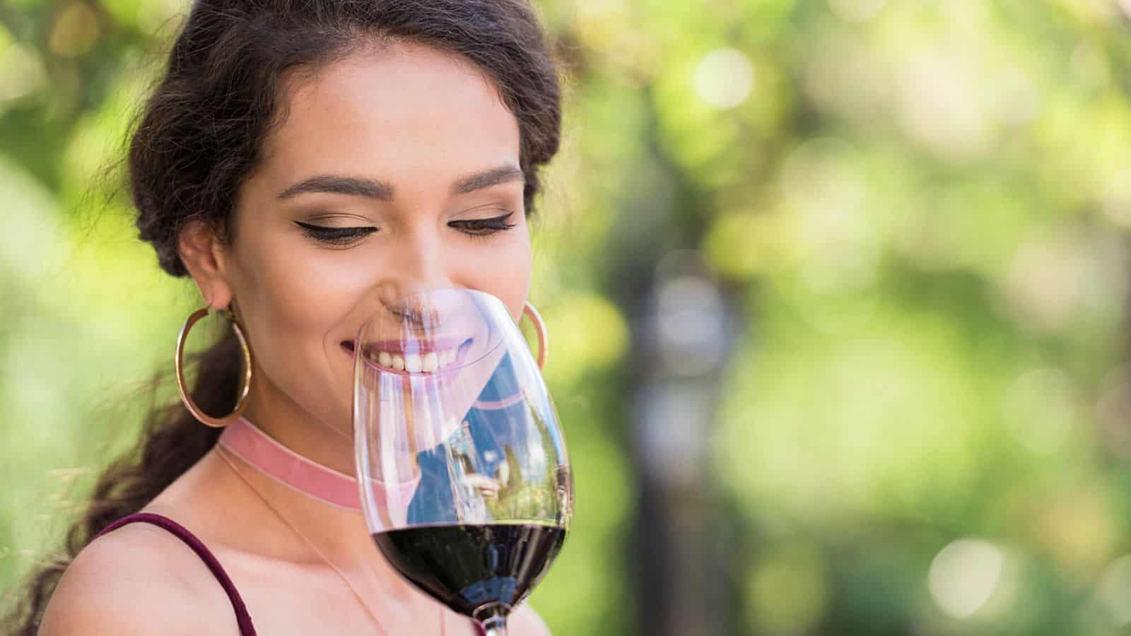 A woman with long dark hair in a ponytail is gently smelling a glass of red wine outdoors. She is wearing hoop earrings and looking down with a slight smile. The background is blurred greenery, suggesting a garden or vineyard setting.