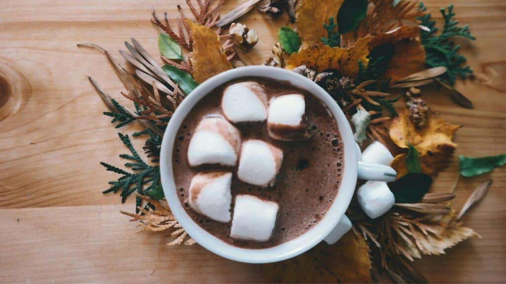 A cup of hot chocolate topped with marshmallows sits on a wooden table. Surrounding the cup are autumn leaves and pinecones, creating a seasonal decoration.