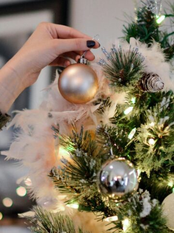 A hand is placing a gold ornament on a decorated Christmas tree. The tree is adorned with various ornaments, including silver and white baubles, and lit with white lights. In the background, a blurred framed picture is visible on the wall.
