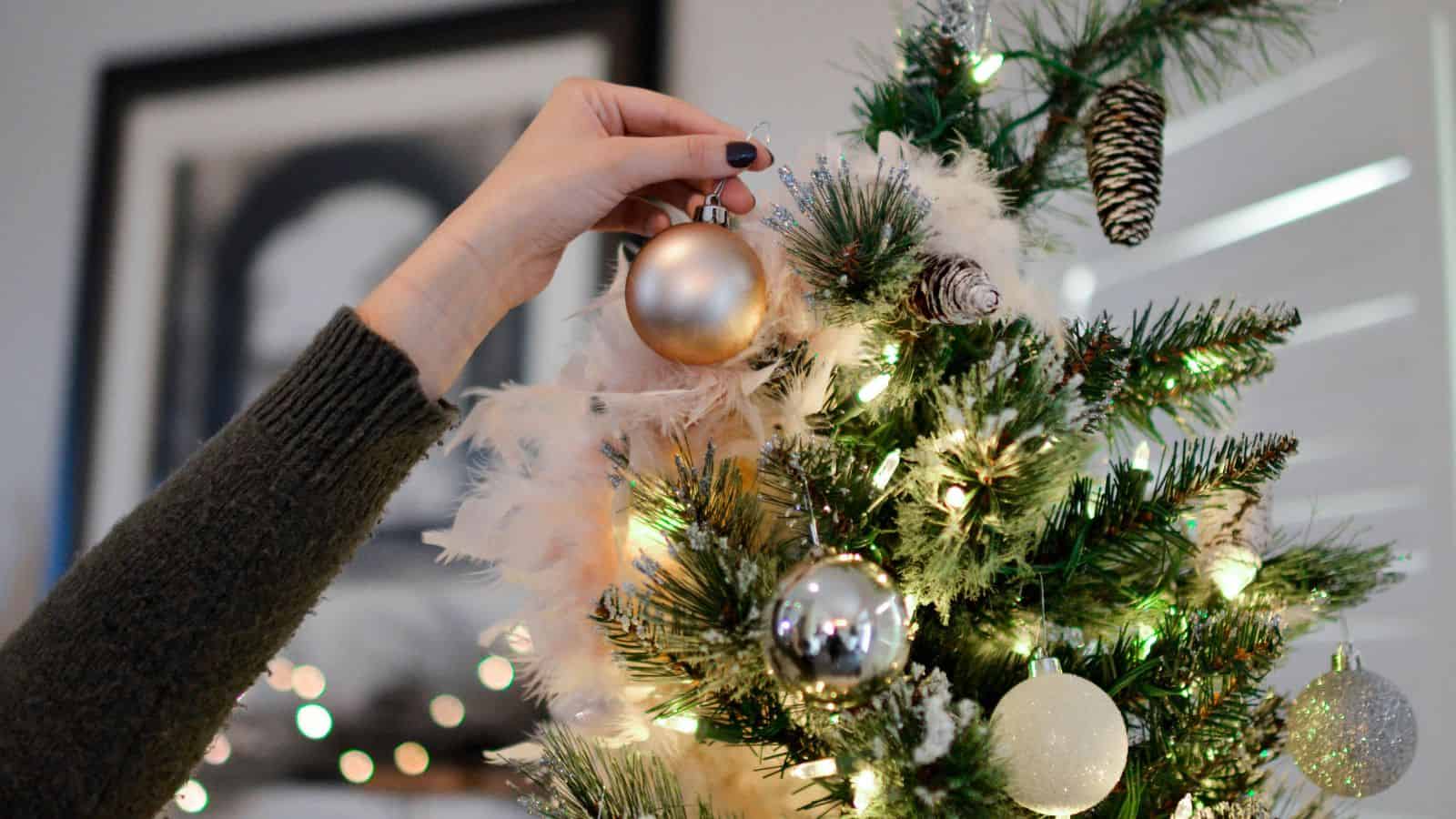 A hand is placing a gold ornament on a decorated Christmas tree. The tree is adorned with various ornaments, including silver and white baubles, and lit with white lights. In the background, a blurred framed picture is visible on the wall.
