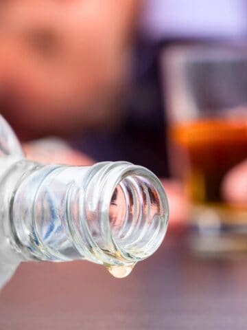 A close-up of an empty glass bottle lying on its side with a drop dripping from the opening. In the blurred background, a person holds a glass of amber-colored liquid. The scene suggests the consumption of alcohol.