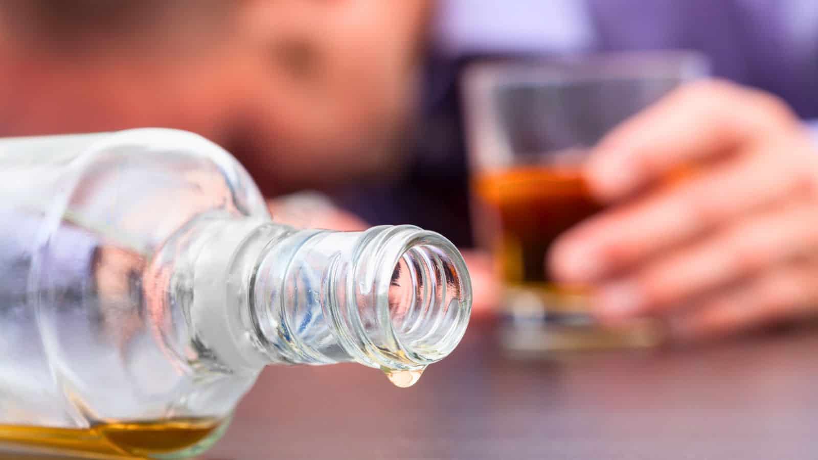 A close-up of an empty glass bottle lying on its side with a drop dripping from the opening. In the blurred background, a person holds a glass of amber-colored liquid. The scene suggests the consumption of alcohol.