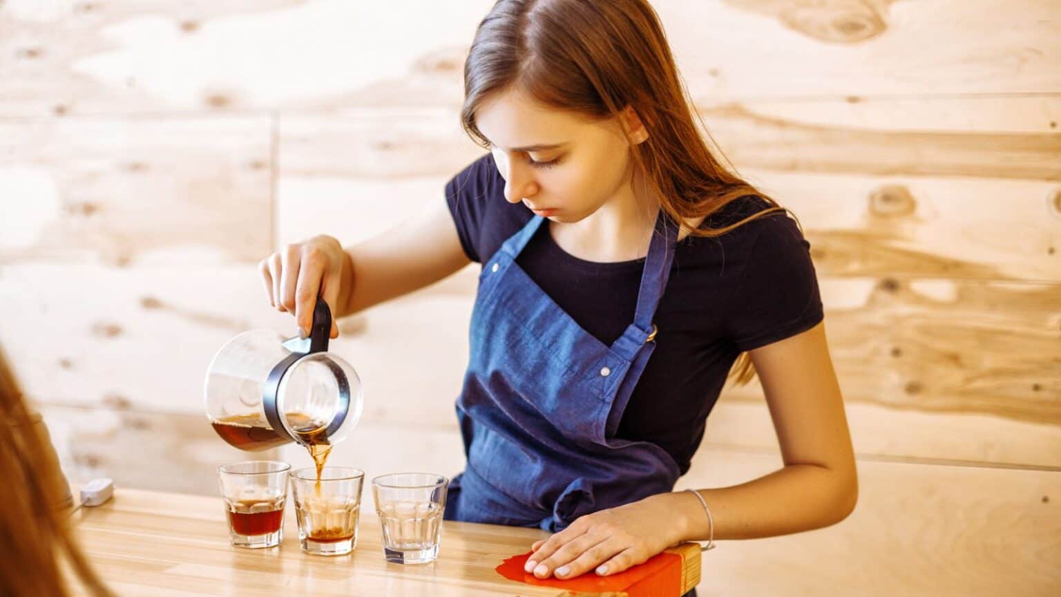 A woman wearing a blue apron is pouring coffee from a pot into three clear glasses on a wooden counter. The background features light wooden panels.