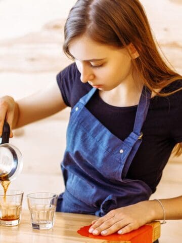 A woman wearing a blue apron is pouring coffee from a pot into three clear glasses on a wooden counter. The background features light wooden panels.