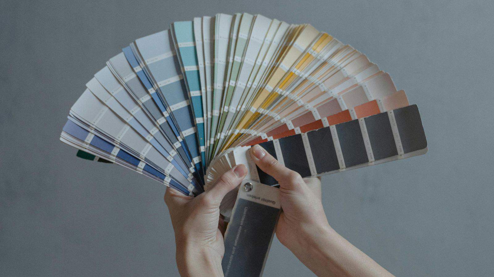 A person is holding a fanned-out set of color swatches against a plain gray background. The swatches display a gradient of colors ranging from blue and green to orange and black.