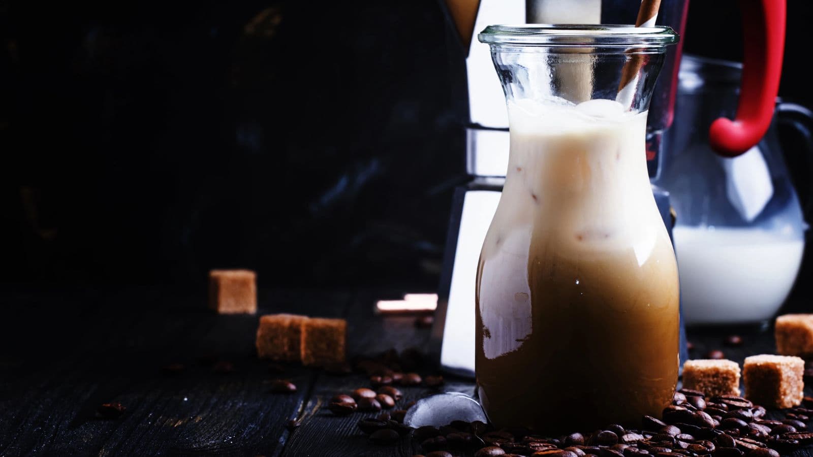 A glass jug filled with iced coffee and milk is on a dark wooden table. Coffee beans and brown sugar cubes are scattered around. In the background, a milk jug and a French press are partially visible.