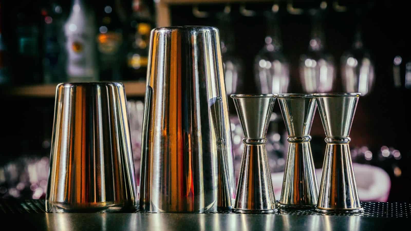 A set of stainless steel cocktail shakers and jiggers are arranged on a bar counter. Various bottles and glassware are blurred in the background.