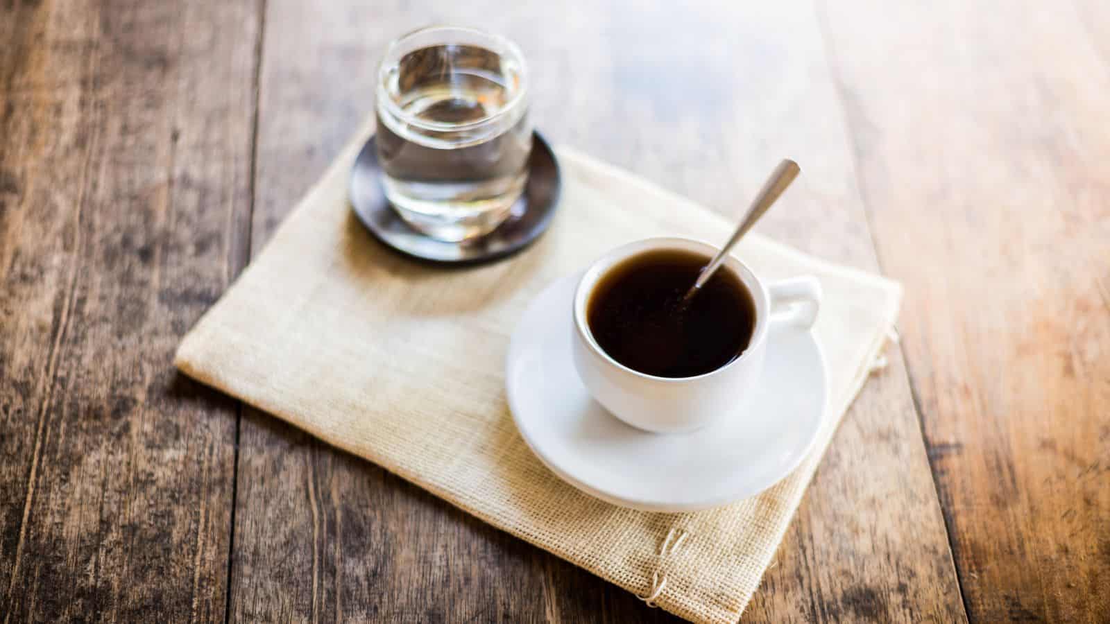 A cup of black coffee with a spoon sits on a saucer next to a glass of water on a small plate. Both are placed on a beige cloth on a wooden table.