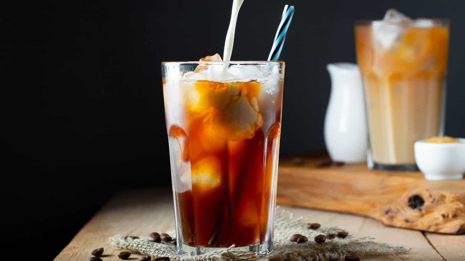 A glass of iced coffee with cream being poured into it, placed on a wooden surface. In the background, there's another glass of iced coffee and a small white pitcher. Coffee beans are scattered on the surface.