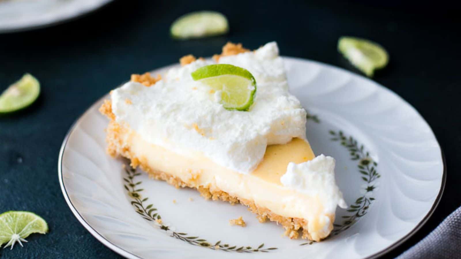 A slice of pie with a crumbly crust, creamy yellow filling, and a layer of whipped topping is on a decorative plate. A lime slice garnishes the top. Lime pieces are scattered on a dark surface around the plate.