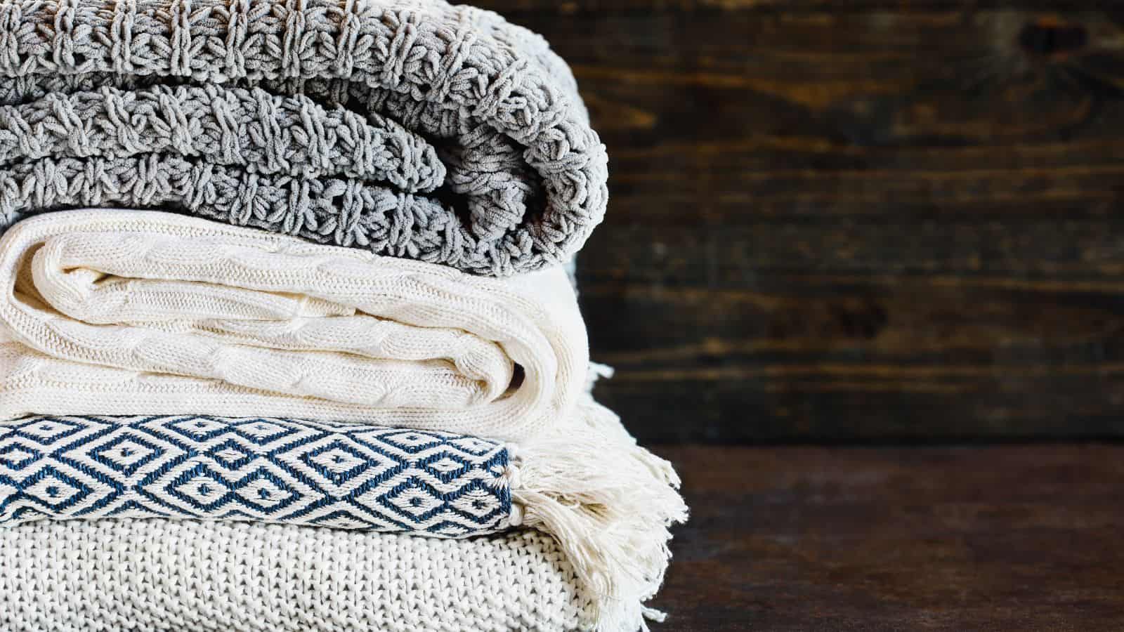 A neatly stacked pile of folded blankets and textiles rests on a wooden surface. The top blanket is textured gray, followed by a cream knitted one, a cream fringed fabric, and a blue and white geometric patterned textile.