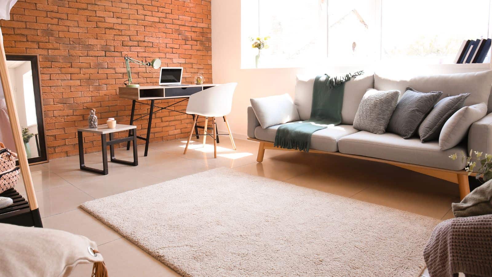 A cozy living room with a modern design features a light gray sofa adorned with gray pillows and a green blanket. A wooden coffee table and a textured rug are placed on the floor. A desk with a chair is situated near a window and a brick wall.