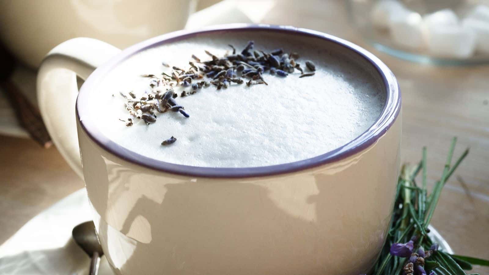 A close-up of a cup filled with a creamy beverage topped with lavender sprigs. The cup has a purple rim and is placed on a table with some herbs and a spoon nearby. Natural light illuminates the scene.
