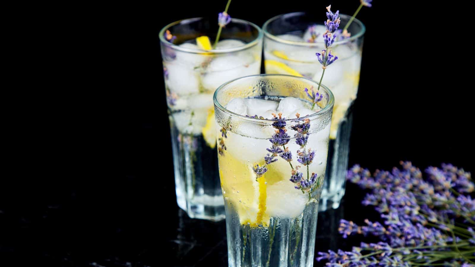 Three tall glasses filled with ice cubes, lemon slices, and lavender sprigs, set against a black background. A small bunch of lavender lays beside the glasses.