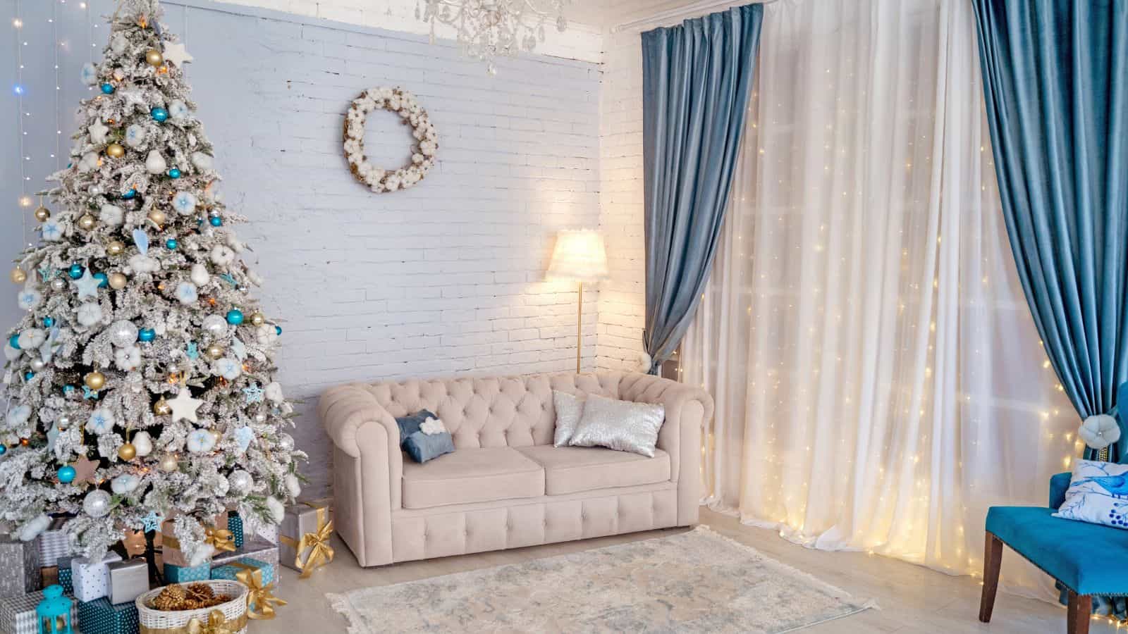 A cozy living room with a decorated Christmas tree, a beige tufted sofa with cushions, and gifts beneath the tree. Blue and white curtains adorned with string lights cover the windows. There is a floor lamp beside the sofa.